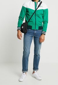 Grön och vit zip-up jacka med randiga ärmslut, ihop med ljusblå jeans och vita sneakers. Liten svart axelremsväska.