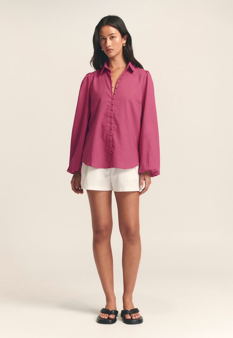 Magenta shirt met lange mouwen, een knoopsluiting aan de voorkant en een kraag, gecombineerd met witte shorts en zwarte sandalen. De stoffen lijken lichtgewichtig en soepel.