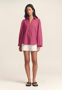 Magenta long-sleeve shirt with a buttoned front and a collar, paired with white shorts and black sandals. Fabrics appear lightweight and smooth.