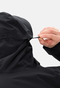 Giacca nera con cappuccio e cordino regolabile. Il materiale sembra liscio e impermeabile. Cuciture dettagliate visibili al collo e alla spalla.