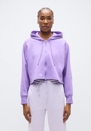 Fialová krátka mikina na zips s kapucňou a šnúrkou, s jemným logom na hrudi a mäkkou, textúrovanou tkaninou. Kombinovaná s svetlofialovými nohavicami.