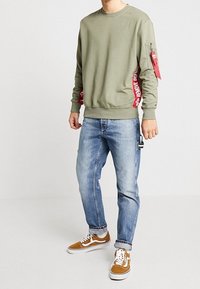 Sudadera de color verde oliva con gráfico de texto rojo en el lateral, puños ajustados y bolsillo con cremallera en la manga; combinada con jeans azul claro y zapatillas marrones.