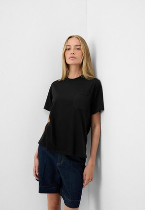 Lyshåret kvinde lænende mod en hvid væg iført en sort kortærmet skjorte med lomme og mørke denimshorts.