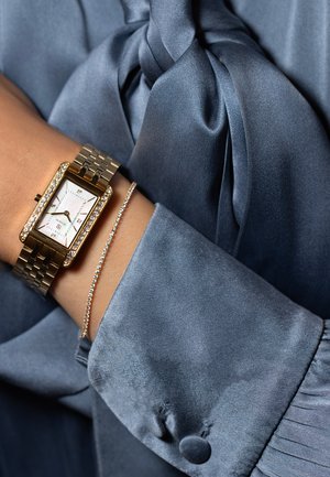 Guldarmbandsur med rektangulär urtavla, diamanthliknande detaljer och ljusfärgad urtavla, ihopparat med ett delikat silverarmband mot en blå satinfabric.