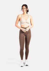 Beige sports-bh med rynket front, parret med brune højtaljede leggings. Hvide sneakers med sorte detaljer fuldender outfittet.