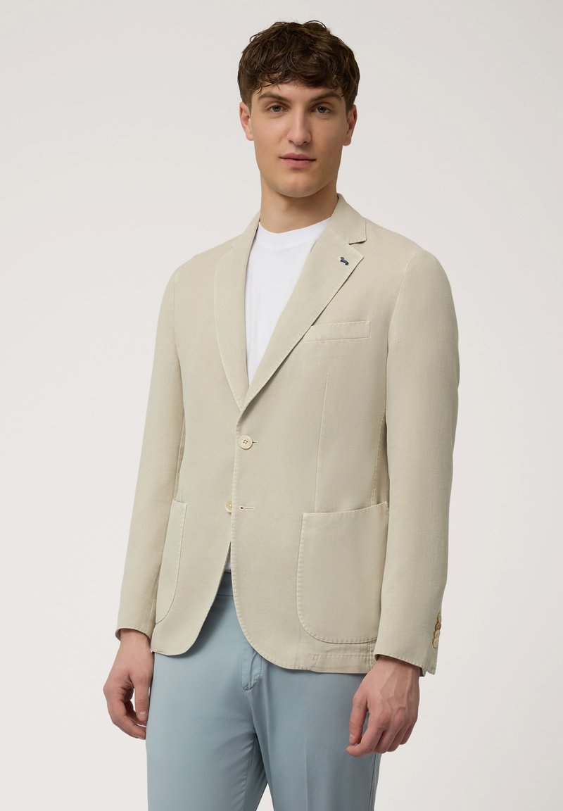 Beige blazer made of textured fabric, featuring a notched lapel, two buttons, and two front pockets. Worn over a white t-shirt.