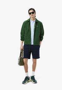 Grön jacka med inredningsmönster, ljusblå polo, marinblå shorts och gröna sneakers med blå och gula accenter. Bär en grön väska.