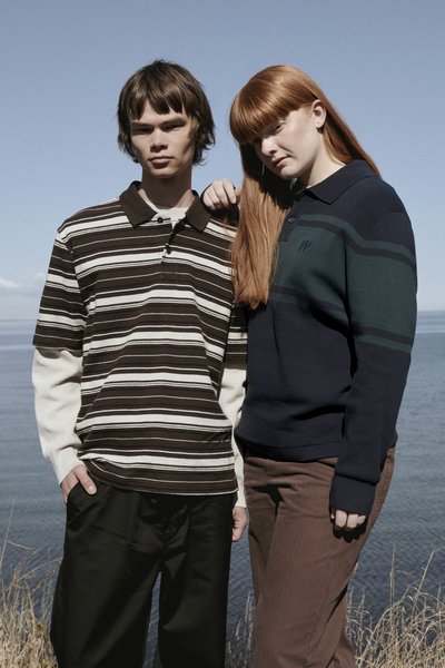 Deux mannequins se tiennent près de l'eau ; l'un porte un polo rayé marron et beige, l'autre un pull bleu marine avec des rayures et un pantalon marron.