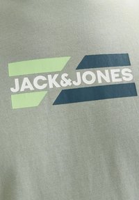Szürke pamut póló, amelyen a "JACK&JONES" felirat fehér színben, zöld és sötétkék geometriai részletekkel látható. Sima tapintású, laza szabású.