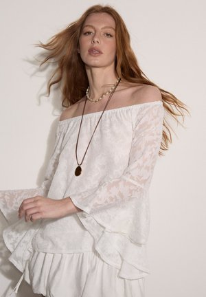 Mujer pelirroja que lleva una blusa blanca de encaje con hombros descubiertos, mangas campana largas y falda en capas, complementada con choker de perlas y collar con colgante.