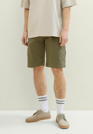 Mężczyzna w beżowych sneakersach, białych skarpetach do kostek w czarne paski, oliwkowych szortach cargo i jasnobeżowej luźnej koszulce.