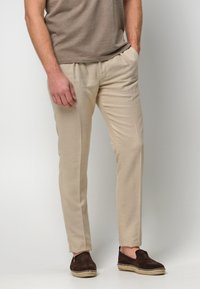 Beżowe lniane spodnie charakteryzują się dopasowanym krojem, bocznymi kieszeniami i fałdami z przodu. Połączone z ciemnobrązowymi butami wsuwanymi z plecionymi detalami.