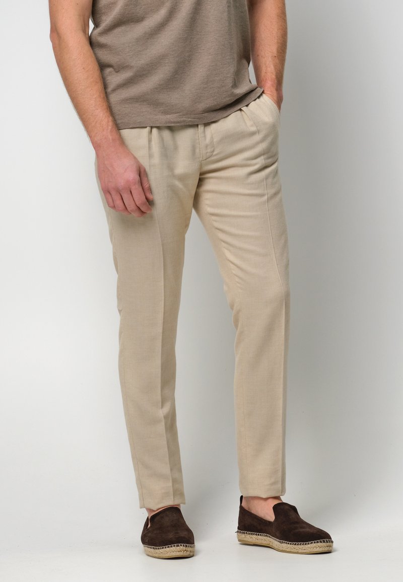 Beżowe lniane spodnie charakteryzują się dopasowanym krojem, bocznymi kieszeniami i fałdami z przodu. Połączone z ciemnobrązowymi butami wsuwanymi z plecionymi detalami.