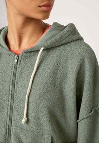 Felpa con zip in morbido verde, caratterizzata da una finitura testurizzata, cappuccio con cordino e accenti crema, e una vestibilità rilassata.