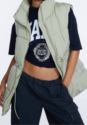 Personne portant une doudoune sans manches vert clair par-dessus un crop top bleu marine avec le logo Yale et un pantalon cargo bleu marine, une main dans une poche.