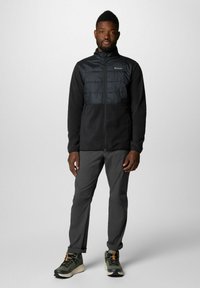 Giacca in pile nera con sezione superiore trapuntata, chiusura frontale con zip, abbinata a pantaloni grigi e scarpe da ginnastica nere con accenti arancioni.