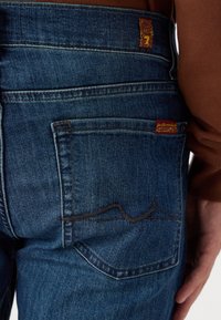 Džínové kalhoty v tmavě modré barvě s výrazným designem zadní kapsy, se zakřiveným švem a hnědým prošíváním, spolu s logovými štítky.