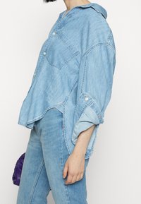 Ljusblå denimskjorta i oversized modell med knappar, en framficka, uppvikta ärmar och en rundad nederkant. Bärs med matchande jeans.