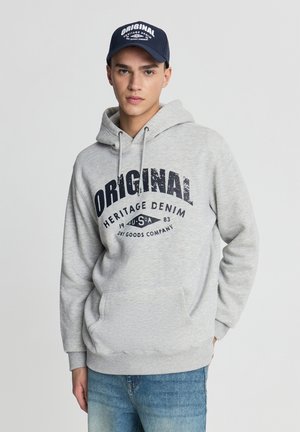 Felpa grigia in cotone con tasca frontale, caratterizzata da una grande scritta blu navy. Abbinata a jeans blu e un berretto blu navy con scritte bianche.