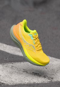 Φωτεινό κίτρινο παπούτσι για τρέξιμο με πορτοκαλί και πράσινες λεπτομέρειες, στον αέρα πάνω από γκρι ασφαλτοστρωμένο δρόμο με λευκές διαγραμμίσεις.
