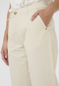 Helle beigefarbene Hose aus Baumwolle mit flachem Vorderteil, Seitentaschen und Knopfverschluss. Glatte Textur mit minimaler Detailgestaltung.