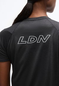 Άτομο με σκούρα μαλλιά δεμένα πίσω, φορώντας μαύρο υφασμάτινο πουκάμισο με λευκά γράμματα "LDN" στο πάνω μέρος της πλάτης.