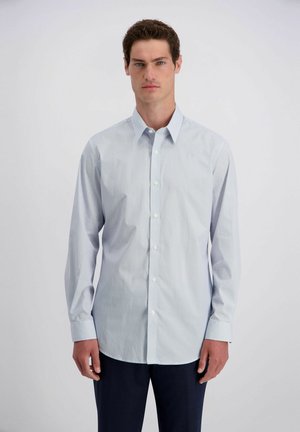 Man met kort krullend haar, gekleed in een lichtblauw patroon overhemd met lange mouwen en een donkere broek, staand tegen een effen witte achtergrond.