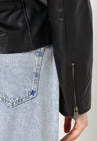 Černá kožená bunda viditelná na rameni, se zipem na manžetě; světle modré džíny denimové s kapsou s malým modrým logem.