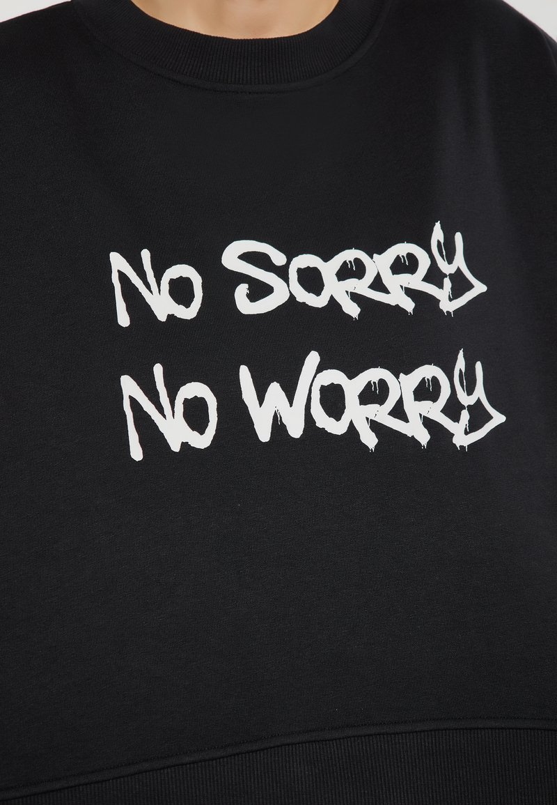 Fekete kapucnis felső, pamutból készült, két sor fehér, graffitis stílusú felirattal: "No Sorry" és "No Worry". Bordás nyakkivágással.