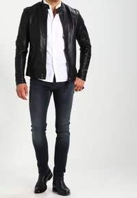 Svart läderjacka med en figursydd design, ståkrage och dragkedjestängning, parat med en vit skjorta och mörka slim-fit jeans.