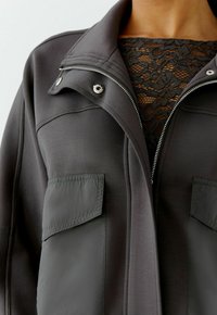 Schwarze Jacke mit Reißverschluss, Druckknöpfen, zwei Vordertaschen und einem strukturierten schwarzen Spitzen-Einsatz. Hergestellt aus einem glatten, flexiblen Stoff.