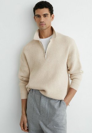 Férfi bézs bordázott, félcipzáras pulóverben, fehér inggel és szürke magas derekú nadrágban, fehér fal előtt áll, kezét zsebében tartva.