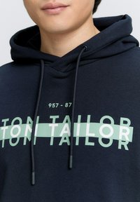 Felpa navy con tasca a marsupio, caratterizzata da una stampa grafica verde menta "TON TAILOR" e numeri. Coulisse con punte in plastica. Texture morbida.