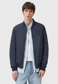 Marine bomberjakke med ribbede mansjetter og krage, glidelås, lett stoff. Båret over en stripete skjorte, parret med lys denim.