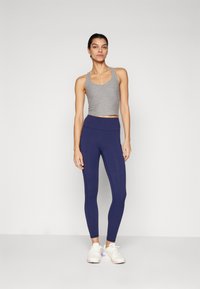 Grå, kort tanktopp kombinert med høyreste navy leggings, som viser en glatt tekstur. Modellen har på seg hvite joggesko med pastellfarger.