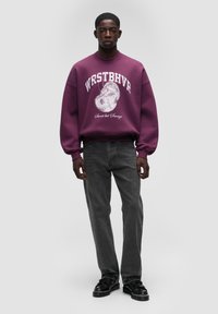 Felpa viola con stampa grafica e testo; vestibilità rilassata, polsini a coste. Abbinata a jeans dritti grigio scuro e scarpe nere.