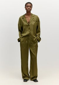 Olivengrünes Satin-Pyjama-Set mit vertikalen schwarzen Streifen, bestehend aus einem Knopfhemd, Sweathosen mit Kordelzug und seitlichen Taschen. Schwarze Schuhe.