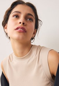 Beige ribbed tank top with a high neck and sleeveless design. Silver hoop earrings accentuate the neckline. Smooth skin texture.