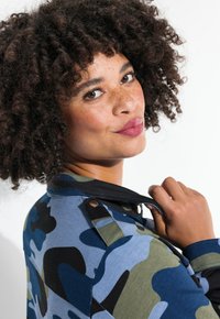 Chaqueta con patrón de camuflaje que presenta tonos azules, verdes y negros; tejido texturizado con un cuello negro y botones en los hombros.