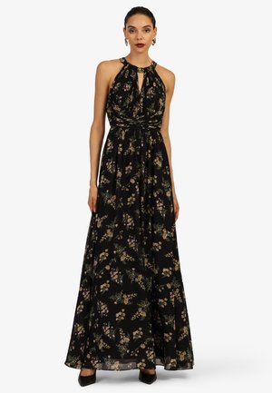 Frau trägt ein schwarzes Maxikleid mit Neckholder und Blumenmuster, tailliert, sowie schwarze spitze High Heels, steht vor weißem Hintergrund.