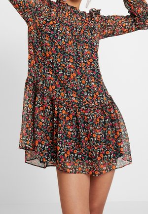 Femme portant une robe noire transparente avec un imprimé floral orange, rouge et blanc, taille cintrée, manches longues bouffantes et un col haut volanté.