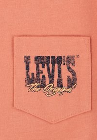 Pomarańczowa kieszeń Levi's z czarnym logo tekstowym "LEVI'S" oraz "the Original" w złotej kursywie. Gładka tekstura bawełny z przeszywaną kieszenią w kształcie sześciokąta.