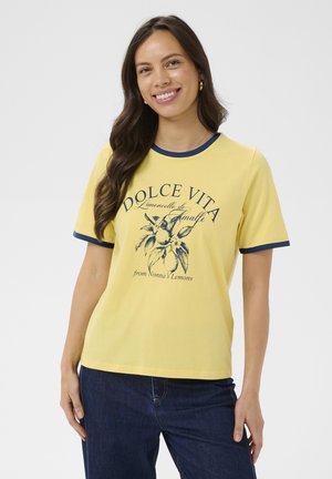 Mladá žena s dlouhými tmavými vlasy se usmívá, má na sobě žluté tričko s grafikou větvičky citronu a nápisy, v kombinaci s tmavě modrými džínami.