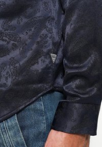 Tumši zils krekls ar teksturētu paisley rakstu, kas iekļauj trīsstūrveida "Guess" logo un gludu, nedaudz mirdzošu auduma apdari.