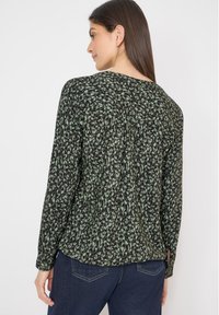 Woman turning sideways wearing a long-sleeve black and green patterned blouse and dark blue jeans against a plain white background.