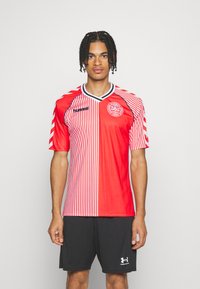 Mies seisoo päällään punavalkoraidallinen jalkapallopaita, jossa on Tanskan jalkapalloliiton logo, ja mustat urheilushortsit.