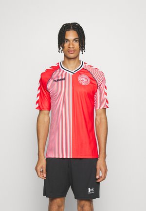 Man staat en draagt een rood-wit gestreept voetbalshirt met het logo van de Deense voetbalbond en zwarte sportbroek.