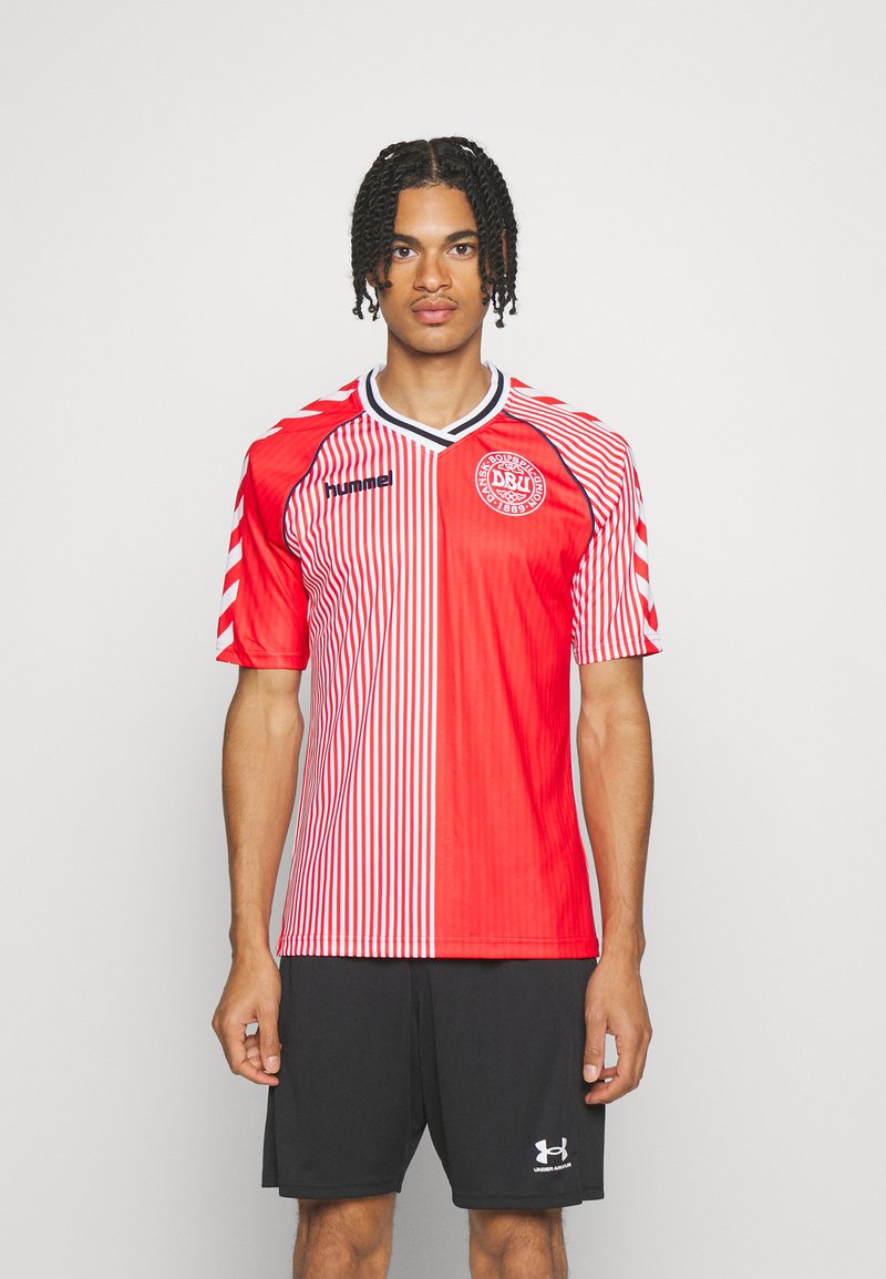 Mies seisoo päällään punavalkoraidallinen jalkapallopaita, jossa on Tanskan jalkapalloliiton logo, ja mustat urheilushortsit.