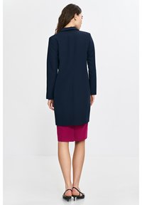 Granatowa marynarka z długimi rękawami, dopasowany krój, z pojedynczym rozcięciem z tyłu, w zestawieniu z fuksjową spódnicą do kolan oraz czarnymi sandałami na paski.