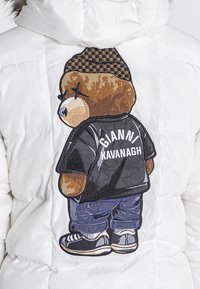 Biała puchowa kurtka z dużym haftem misia na plecach, noszącego czarną koszulę z napisem "GIANNI KAVANAGH" i niebieskie spodnie.